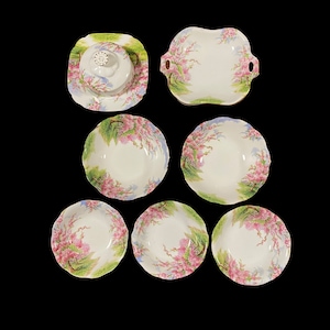 Op de afbeelding: Een set van zeven witte porseleinen kommen met een bloemenmotief. De kommen zijn in verschillende maten en vormen, waaronder een botervloot met deksel. De kommen hebben een delicaat bloemenmotief met roze en groene accenten.