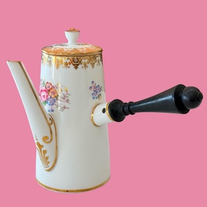 May include: A white porcelain chocolate pot featuring a cylindrical body, a long angled spout, and a black wooden side handle. The exterior is decorated with hand-painted floral motifs in pink, blue, and yellow, accented by ornate gold trim and gilding.