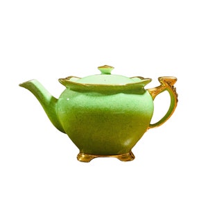 May include: A green teapot with gold accents and a lid. The teapot has a round body and a spout on one side. The handle is on the other side and is shaped like a curled leaf. The teapot is sitting on a white background.