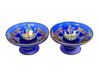 Op de afbeelding: Twee kobaltblauwe glazen dessertkommen met voetstukken. Elke kom heeft een gouden rand en sierlijke gouden en bloemmotieven. De binnenkant van elke kom heeft een geschilderde scène van figuren in historische kleding.