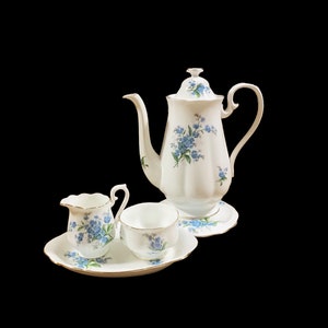 Może przedstawiać: Biały porcelanowy zestaw do herbaty z kwiatowym wzorem z niebieskimi niezapominajkami. Zestaw zawiera czajnik, dzbanek do śmietanki, cukiernicę i pasujący spodek.