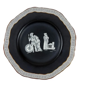 Puede incluir: Plato de cerámica negra con borde festoneado y borde blanco con diseño de greca. El plato presenta un fondo negro con ilustraciones blancas de figuras clásicas y un edificio.