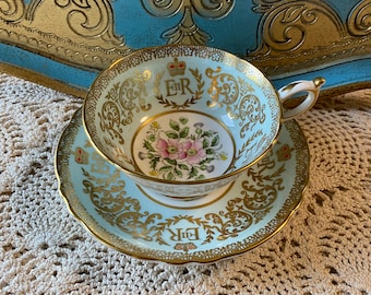 May include: A light blue teacup and saucer with gold trim and a floral design. The cup features a crown and the letters "E.R." in gold.