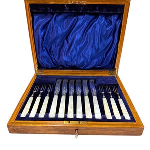 May include: A wooden box with a blue velvet lining, containing a set of 12 silver-plated knives and forks with mother-of-pearl handles.