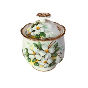 Peut inclure: Un pot en céramique blanc avec un couvercle, orné d'un motif floral de fleurs blanches à centres jaunes et de feuilles vertes. Le pot et le couvercle sont bordés de dorures. Le pot est probablement destiné à conserver du sucre ou d'autres petits articles.