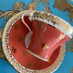 May include: A coral-colored teacup and saucer set with gold trim and a floral pattern. The cup and saucer are decorated with delicate gold leaves and flowers.