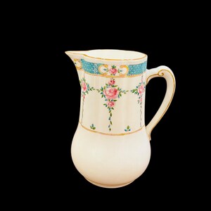 May include: A white ceramic pitcher with a rounded body and a handle. The pitcher is decorated with pink roses and green leaves, with a blue and gold trim around the top. The pitcher has a spout for pouring.