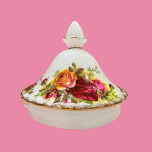 Könnte beinhalten: Eine weiße Porzellan-Servierschüssel mit Deckel und einem Rosenmuster in Rot-, Gelb- und Rosatönen. Der Rand und der Knauf sind goldfarben verziert. Die Schüssel steht vor einem rosa Hintergrund.