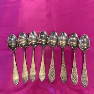 May include: Set of eight silver spoons with ornate designs on a pink fabric background.
