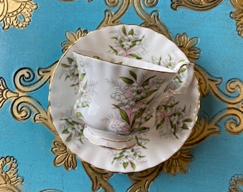May include: A white teacup and saucer with a delicate floral pattern featuring pink and white lilies of the valley. The cup and saucer are on a blue and gold tray.