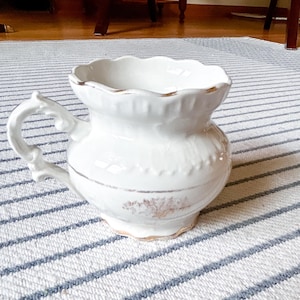 May include: A white ceramic chamber pot featuring a scalloped rim, a decorative handle, and a subtle gold-toned floral pattern around the center. The base and the upper edge are accented with a thin gold trim, giving the piece a defined appearance.