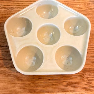 May include: A cream-colored ceramic egg holder with six circular indentations. The holder has a pentagon shape and sits on a wooden surface. The egg holder is designed to hold six eggs.
