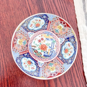 May include: A decorative ceramic plate with a detailed floral design. The plate features a central image of flowers and bamboo, surrounded by a blue, red, and gold patterned border. The plate is round and has a gold rim.