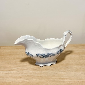 May include: White ceramic gravy boat with a scalloped rim and a handle. The boat features a blue floral pattern around the base and is set on a wooden surface. The gravy boat is a classic piece for serving sauces and gravies.