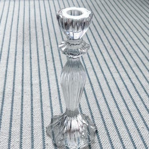 May include: A clear, glass candlestick holder with a fluted design. The candlestick has a wide, star-shaped base, a slender, ribbed stem, and a ruffled midsection. The top has a circular opening for a candle.