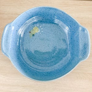 May include: A round, light blue ceramic dish with two handles. The dish has a speckled texture and a painted design of a yellow and green sun. The dish is suitable for serving food.