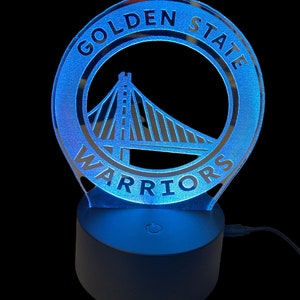Peut inclure: Une veilleuse avec le logo des Golden State Warriors en bleu et argent, avec une base dotée d'un interrupteur. Le logo représente le Golden Gate Bridge.