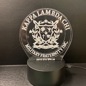 May include: A black and white acrylic night light with a Kappa Lambda Chi Military Fraternity logo. The logo features a shield with a cross, a star, and a wreath. The text "Zeta Eta Spr 24" is printed below the logo.