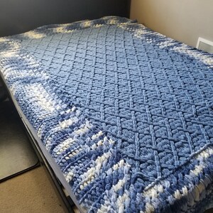 Puede incluir: Una manta de crochet azul y blanca con un patr&oacute;n de diamante. La manta est&aacute; en una cama con una cabecera negra.
