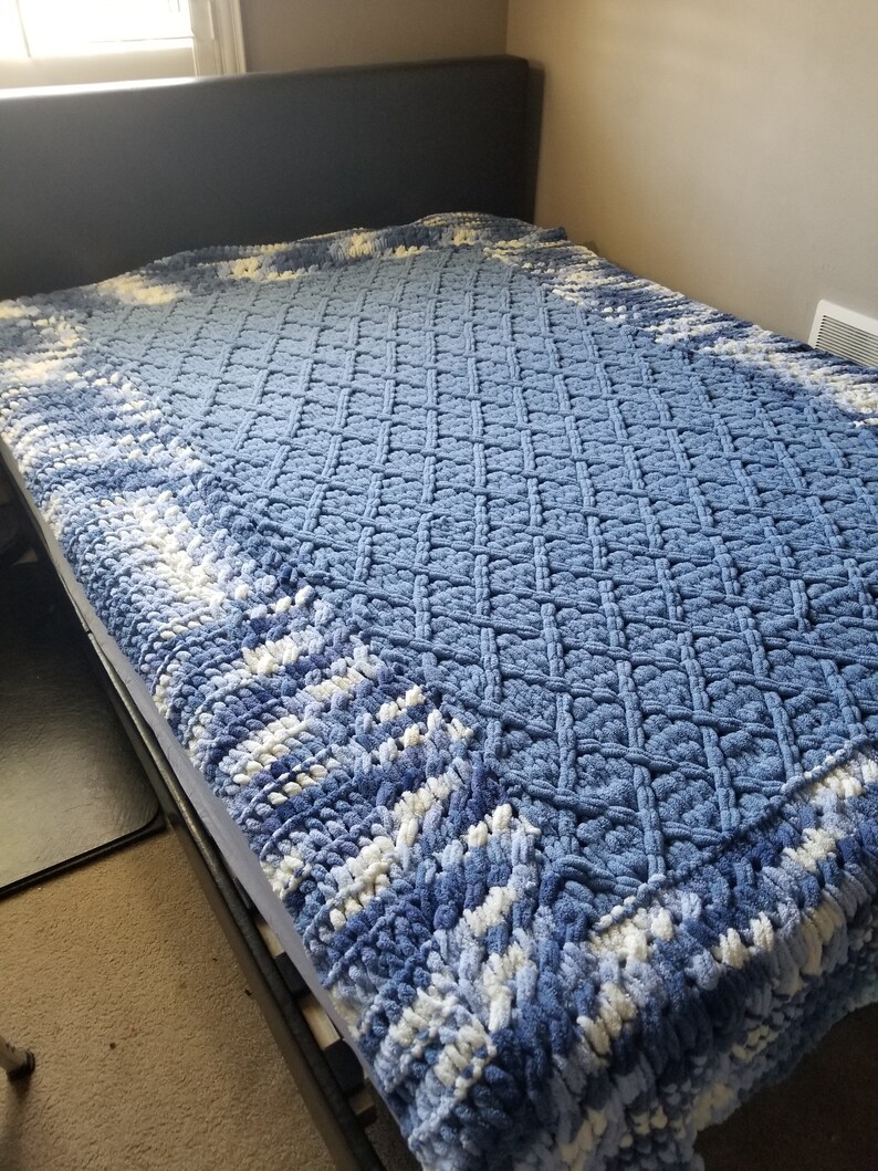 Puede incluir: Una manta de crochet azul y blanca con un patr&oacute;n de diamante. La manta est&aacute; en una cama con una cabecera negra.