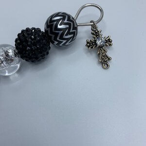 以下が含まれることがあります： 銀と黒のビーズブレスレット。中央にクリアなラインストーンがついた十字架の形をした銀のチャームが付いています。ブレスレットは銀色の留め具が付いています。