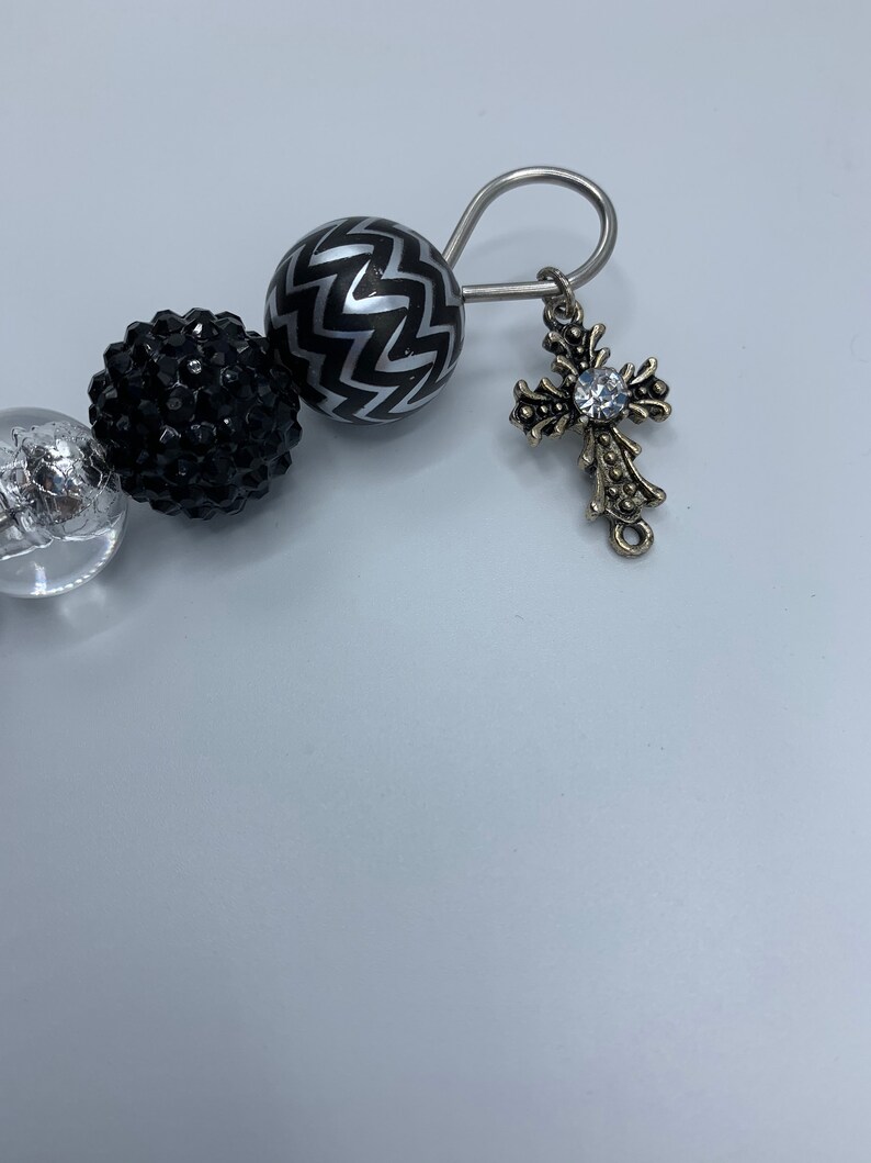 以下が含まれることがあります： 銀と黒のビーズブレスレット。中央にクリアなラインストーンがついた十字架の形をした銀のチャームが付いています。ブレスレットは銀色の留め具が付いています。