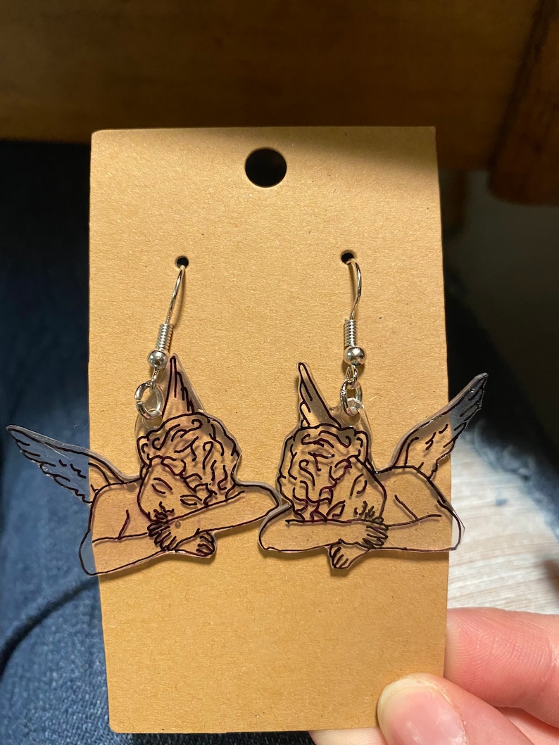 May include: A pair of clear acrylic earrings featuring a black line drawing of a sleeping angel with wings. The earrings have silver hooks.