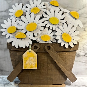 May include: A wooden basket filled with white daisies with yellow centers. The basket has a brown wooden base and two handles. A yellow tag with the text "freshly picked daisies" is attached to the handle.