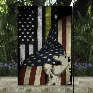 May include: A garden flag featuring a distressed American flag design with a fist clenching the flag. The flag has a black and yellow stripe, and the stars and stripes are visible. The flag is displayed in a garden setting.