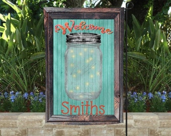 May include: A rectangular garden flag with a wooden frame features a mason jar filled with glowing lights against a teal background. The word "Welcome" arches across the top, and "Smiths" is printed at the bottom in red.