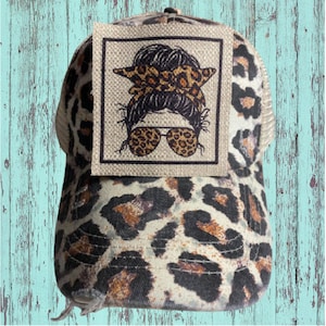 May include: A leopard print trucker hat with a brown and black patch featuring a cartoon illustration of a woman with a messy bun, leopard print headband, and leopard print sunglasses.