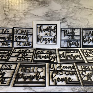 May include: A collection of 12 black wooden signs with various phrases, including "Love Begins at Home", "Land of the Free", "We Are Family", "Welcome to Our Home", and "Hello Spring".