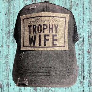 May include: Black mesh trucker hat with a distressed look. The hat has a brown patch with the text "participation trophy wife" in black.