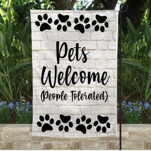 May include: A white garden flag featuring black paw prints and the text "Pets Welcome (People Tolerated)" in black script. The background has a white brick effect. The flag is displayed outdoors.