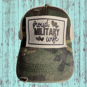 May include: A camouflage trucker hat with a beige patch that says "proud military wife" with glitter hearts.
