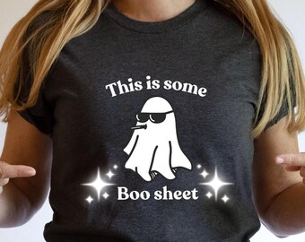 May include: Dark gray t-shirt with the text "This is some Boo sheet" and a white graphic of a ghost wearing sunglasses. The shirt is a crew neck and is being pointed at by a person.