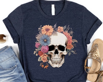 May include: Navy blue t-shirt featuring a skull design adorned with colorful flowers. The graphic includes a skull with a floral crown in shades of pink, orange, blue, and yellow. The shirt is paired with denim shorts and gold sandals.
