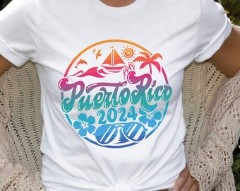 May include: White t-shirt with a colorful Puerto Rico 2024 graphic. The design features a sailboat, palm trees, sunglasses, and hibiscus flowers in a circular arrangement. The shirt is paired with blue jeans and a beige cardigan.