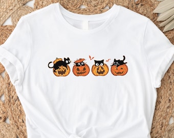 May include: White t-shirt featuring a Halloween design. The graphic shows four orange jack-o'-lanterns with black cats sitting on top. The cats have various expressions. The shirt is tied at the waist.