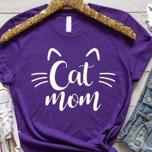 May include: A vibrant purple t-shirt featuring the text "Cat Mom" in white, designed with cat-like features. The shirt is presented on a gold sequin hanger, with white trainers and denim shorts visible.