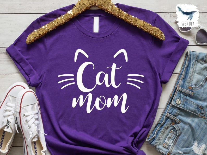 May include: A vibrant purple t-shirt featuring the text "Cat Mom" in white, designed with cat-like features. The shirt is presented on a gold sequin hanger, with white trainers and denim shorts visible.