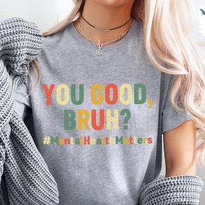 以下が含まれることがあります： カラフルな文字で「YOU GOOD BRUH？」と書かれたグレーのTシャツ。メインテキストの下には「#Mental Health Matters」というフレーズがあります。シャツは半袖で、クルーネックです。