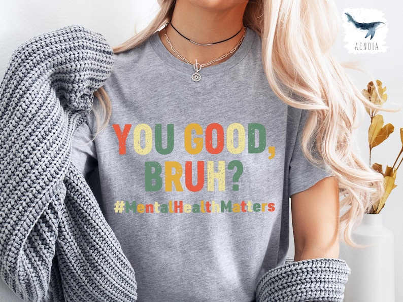 以下が含まれることがあります： カラフルな文字で「YOU GOOD BRUH？」と書かれたグレーのTシャツ。メインテキストの下には「#Mental Health Matters」というフレーズがあります。シャツは半袖で、クルーネックです。