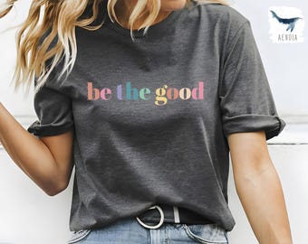 May include: A dark gray t-shirt with the text "be the good" printed in rainbow colors.