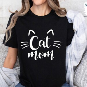 May include: Black t-shirt featuring the words "Cat mom" in white, with cat ear and whisker graphics. The design is in a casual, handwritten style. A comfortable top for cat owners, perfect for everyday wear.