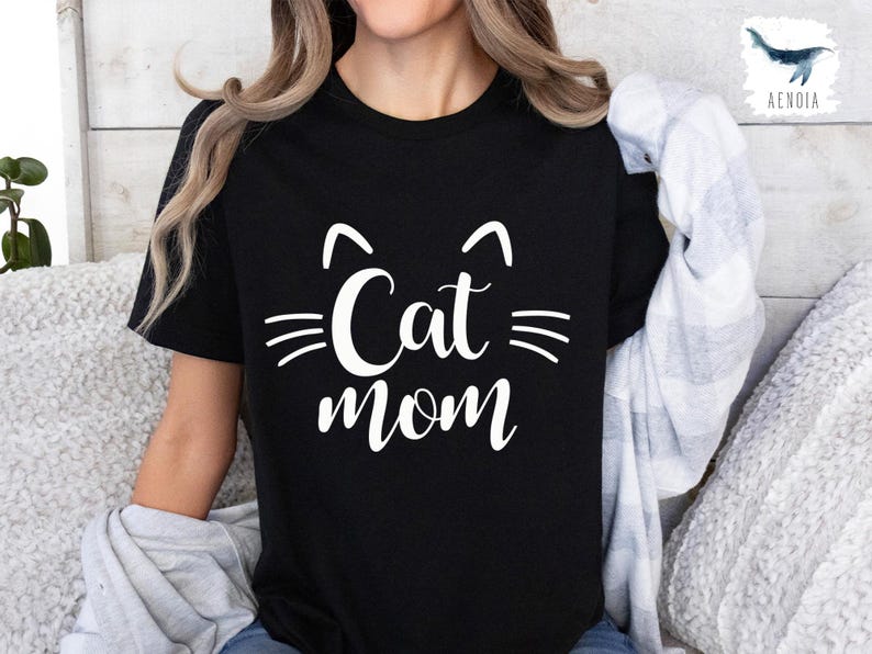 May include: Black t-shirt featuring the words "Cat mom" in white, with cat ear and whisker graphics. The design is in a casual, handwritten style. A comfortable top for cat owners, perfect for everyday wear.