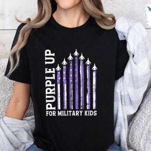 Op de afbeelding: Zwart T-shirt met een "Purple Up for Military Kids"-afbeelding. De afbeelding toont een Amerikaanse vlag in paars en wit met vliegtuigen in formatie.