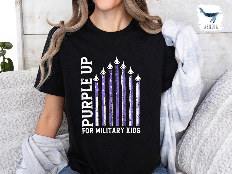 Op de afbeelding: Zwart T-shirt met een "Purple Up for Military Kids"-afbeelding. De afbeelding toont een Amerikaanse vlag in paars en wit met vliegtuigen in formatie.
