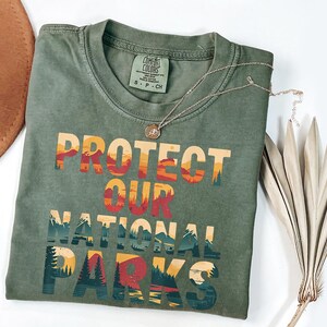 May include: Sage green t-shirt featuring the text "PROTECT OUR NATIONAL PARKS" in a layered design with orange, red, and blue hues. A silver necklace and a dried plant are also visible. A brown hat is in the background.