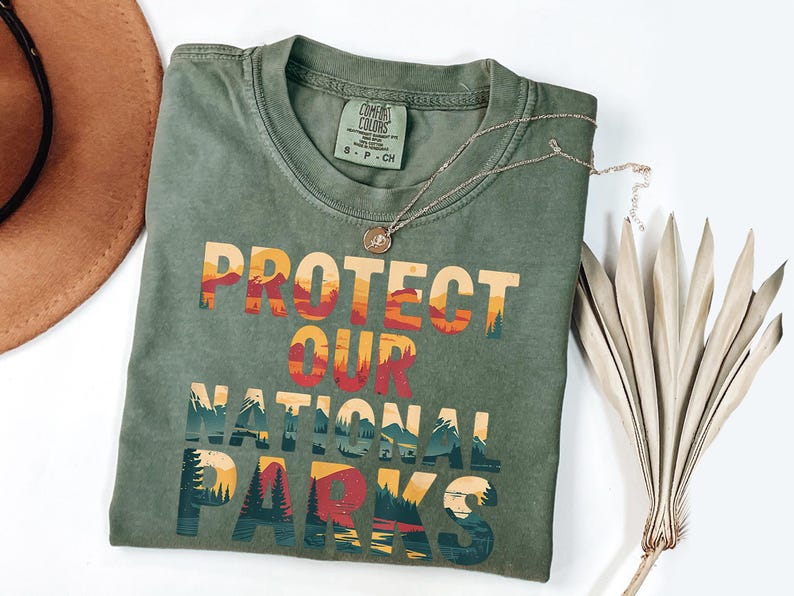 May include: Sage green t-shirt featuring the text "PROTECT OUR NATIONAL PARKS" in a layered design with orange, red, and blue hues. A silver necklace and a dried plant are also visible. A brown hat is in the background.